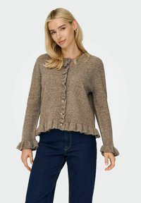 Braune, strukturierte Strickjacke mit einem Rüschensaum und -bündchen, die eine knopfbare Vorderseite hat. Trägt man mit dunkelblauen Jeans.