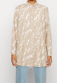 Chemise beige à boutons avec des motifs abstraits blancs, manches longues, col classique et coupe décontractée, associée à un jean bleu clair.