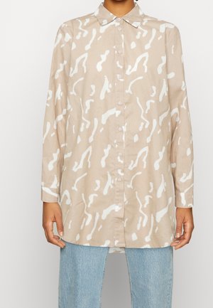 Chemise beige à boutons avec des motifs abstraits blancs, manches longues, col classique et coupe décontractée, associée à un jean bleu clair.