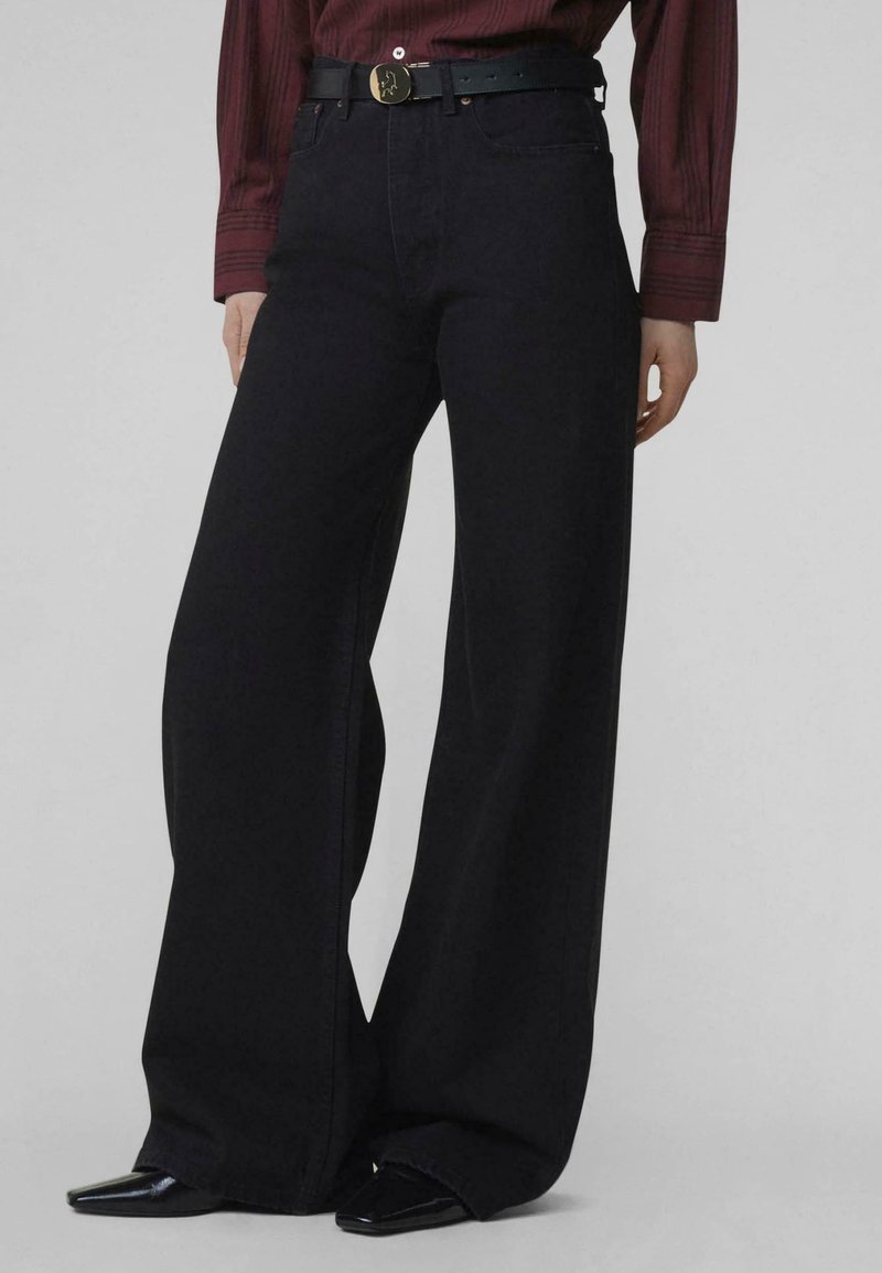 Une personne porte un pantalon noir à jambes larges, une ceinture noire avec une boucle dorée, des chaussures noires pointues, et une chemise rayée rouge foncé à manches longues.
