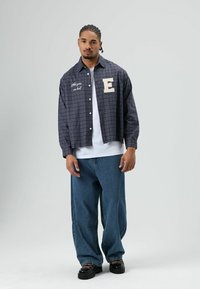 Informeel geruit overhemd in marineblauw en beige met geborduurde details, gecombineerd met losse blauwe spijkerbroek en zwarte schoenen.