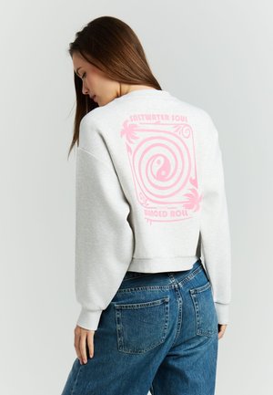 Vrouw draagt een lichtgrijze trui met een roze spiraal- en palmbomenontwerp en de tekst "Saltwater Soul Balanced Roll" op de achterkant, gecombineerd met een blauwe jeans.