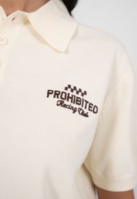 Polo blanc avec un col beige et le texte « PROHIBITED Racing Club » brodé en marron, accompagné d'un motif de drapeau à damier.