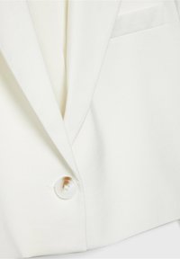 Blazer blanc présentant une coupe structurée, fermeture à un bouton, revers nets et un détail de poche minimaliste. Texture de tissu lisse.