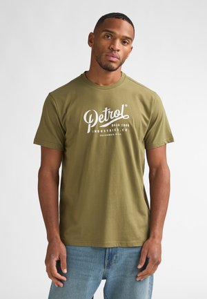 Man met kort haar en baard, gekleed in een olijfgroen "Petrol Industries" t-shirt en blauwe jeans, staand tegen een effen lichte achtergrond.