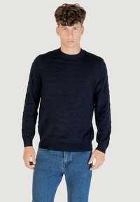 Dunkelblauer Strickpullover mit Wellenmuster; runder Ausschnitt und gerippte Bündchen; kombiniert mit blauen Jeans, die einen strukturierten Stoff zeigen.
