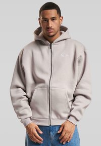 Lys grå zip-up hettegenser laget av myk stoff har en frontlomme og ribbekantede mansjetter, med en subtil brodert logo på brystet.