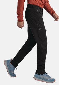 Schwarze Outdoor-Hosen mit schlanker Passform, ausgestattet mit Reißverschlusstaschen und Logo-Akzenten. Getragen mit hellblauen Trailschuhen mit orangefarbenen Details.