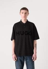 Juodas polo marškinėliai su trimis mygtukais ir pakeltu 'HUGO' logotipu priekyje. Dėvimi su šviesiai mėlynomis džinsinėmis kelnėmis. Paprastas, kasdieniškas dizainas.