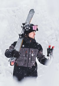 Équipement de ski et bâtons tenus par une personne portant une veste noire et grise avec des accents orange, un casque noir et des lunettes roses, dans un terrain enneigé.
