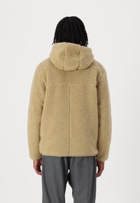 Person mit schwarzen geflochtenen Haaren, die eine beigefarbene Sherpa-Fleece-Jacke mit Kapuze und grauen Hosen trägt, mit dem Rücken zu einer weißen Hintergrund.