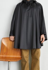 Svart vattentät poncho med huva, som har en hög krage och vida ärmar, kombinerad med khakifärgade byxor, stående bredvid en trästol.