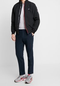 Czarna kurtka bomberka z czerwono-czarnym zamkiem błyskawicznym, szary sweter, granatowe spodnie oraz szare sneakersy z czerwonymi akcentami, stojące na neutralnym tle.