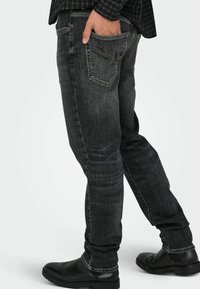 Juodos spalvos džinsai iš dino audinio, siaurėjantys prie kojų, su klasikiniu penkių kišenių dizainu ir subtiliu išblukimu. Dėvimi su juodais odiniais batais.