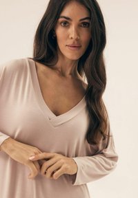 Kobieta z długimi ciemnymi włosami, ubrana w luźną, jasnoróżową bluzkę z dekoltem w kształcie litery V, delikatnie trzymająca ręce razem na tle neutralnym.