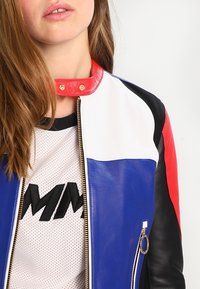 Jeune femme aux longs cheveux portant une veste en cuir color-block blanche, bleue, rouge et noire avec une fermeture éclair à l'avant et un col à pression.