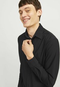 Camicia nera a maniche lunghe realizzata in tessuto morbido, con colletto e polsini allacciabili, mostrata su un modello che regola il colletto.