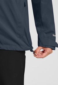 Veste imperméable marine avec un cordon de serrage à l'ourlet, des poignets ajustables et une finition lisse et mate. Positionnée sur un fond neutre.