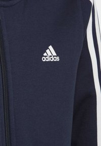 Chaqueta de cremallera azul marino hecha de una mezcla de algodón, con acentos en blanco con tres rayas en las mangas y un pequeño logo blanco de Adidas en el pecho.