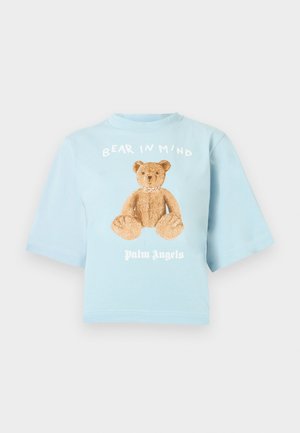 Világoskék rövidített póló rövid ujjakkal, barna mackó grafikával, rajta a "BEAR IN MIND" felirat és alatta a "Palm Angels" szöveg.