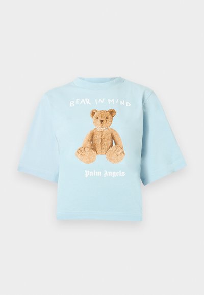 Ανοικτό μπλε κοντομάνικη κοντή μπλούζα, με κοντά μανίκια, που έχει γραφικό του αρκουδάκι σε καφέ χρώμα με το κείμενο "BEAR IN MIND" και "Palm Angels" από κάτω.