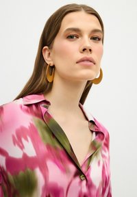 Camisa corta de manga con flores rosas y verdes, con cuello y detalle de botones, combinada con grandes pendientes de aro amarillos. Textura suave y sedosa.