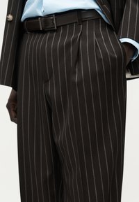 Μαύρο σακάκι με ρίγες, παντελόνι με πτυχώσεις μπροστά, ζώνη από μαύρο δέρμα και μανίκι σακακιού που ταιριάζει πάνω σε μανσέτα ανοιχτό μπλε πουκαμίσου.