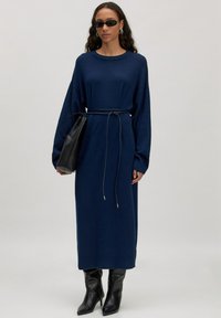 Langärmliges, marineblaues Strickkleid mit lockerer Passform, in der Taille mit einem dünnen Gürtel tailliert; kombiniert mit schwarzen knielangen Stiefeln mit Absatz.