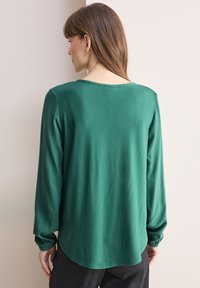 Maglietta verde a maniche lunghe con vestibilità rilassata, caratterizzata da un colletto rotondo, texture a coste e polsini arricciati. Tessuto liscio e leggero.