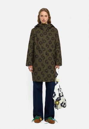 Kobieta nosząca zielony płaszcz z kapturem w kwiatowy wzór, ciemnoniebieskie spodnie, brązowe buty z zielonymi sznurowadłami, trzymająca białą torbę z ciemnym kwiatowym nadrukiem.