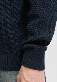 Pull en maille bleu marine avec un motif de câble texturé et des poignets côtelés, mettant en valeur un tissu doux avec un aspect épais.