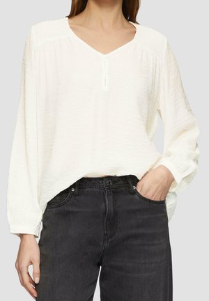 Kvinde iført en løs hvid langærmet bluse, stoppet ned i højtaljede sorte denimjeans med den ene hånd i lommen.