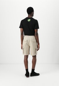 Black t-shirt with green circular logo, paired with beige cargo shorts featuring multiple pockets and an elastic waistband, worn with black socks and sneakers.