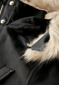 Veste noire avec une capuche en fourrure. Elle présente un tissu lisse, des boutons en métal doré et une accent en fourrure texturée. Comprend des détails de poches à l'avant.