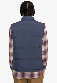 Navy puffer vest, quilted design, high collar, and sleeveless. Worn over a multicolored plaid shirt. Smooth texture, casual style.
