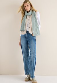 Écharpe côtelée bleu clair drapée sur une veste polaire beige, portée sur une chemise blanche à manches longues, associée à un jean large bleu et des baskets beiges.