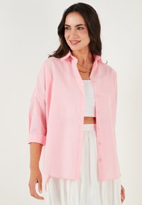 Light pink button-up shirt with a collar, roll-up sleeves, and a relaxed fit. Made of soft fabric, featuring subtle stitching details.
