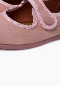 Zapatos de ante rosa con puntera redondeada, tirante elástico y bordes cosidos. Presentan una textura suave y un detalle de botón en la correa.