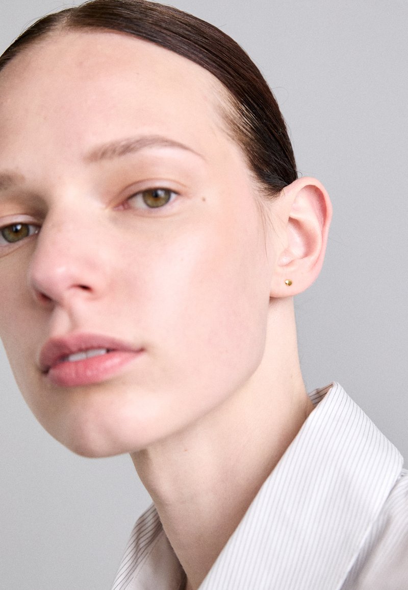 Pogled od blizu na osobu s svetlo kožo, koja nosi male zlatne uhane na klik. Značajke uključuju ravnu kosu, ovratnik na prugice i neutralan izraz lica.