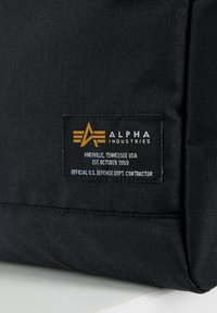 Bolsa de tela negra con una superficie texturizada y una etiqueta destacada con letras doradas y blancas, que indica "Alpha Industries" y otros detalles.
