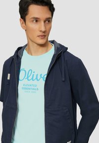 Tumed zip-up jakk, millel on kapuuts ja kaks esikotti, kantud helesinise särgi peale, millel on kiri "Olive Elevated Essentials."
