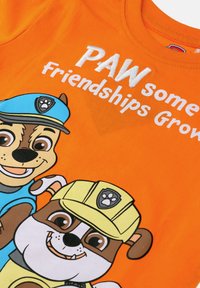 Orange bomulds T-shirt med tegneseriehunde i blå og gule uniformer. Teksten siger: "PAW some Friendships Grow" i hvid skrift.