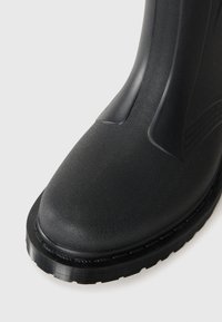 Μαύρη καουτσούκ μπότες με υφασμάτινη εξωτερική επιφάνεια, στρογγυλή μύτη και παχιά, ραβδωτή σόλα. Διαθέτει λεία επιφάνεια στον άξονα για επιπλέον στήριξη.