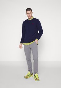 Maglione blu navy sopra una camicia verde con colletto, pantaloni grigi e scarpe da ginnastica multicolori. Presenta un logo rosso sul maglione e materiali testurizzati.