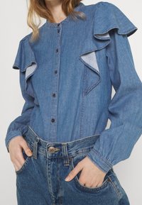 Jeanshemd mit Rüschenschlaufen, hellblauer Farbe, Knopfleiste und Manschetten. Kombiniert mit hoch taillierten, hellblauen Jeans.