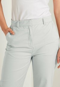 Pantaloni in tessuto elastico grigio chiaro con vita piatta, tasche laterali e superficie liscia, parzialmente infilati sotto una maglietta bianca.