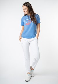 Blauwe katoenen t-shirt met grafische tekst en witte jeans. Model draagt witte sneakers met lichtblauwe accenten. Casual, slim-fit stijl.