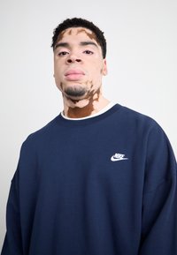 Sudadera azul marino con cuello redondo y logo blanco de Nike. Presenta hombros caídos y un ajuste relajado. Tejido suave y texturizado.