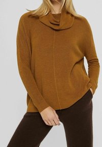 Pull jaune moutarde côtelé avec un col bénitier, coupe ample et poches latérales. Associé à un pantalon marron foncé en tissu texturé.