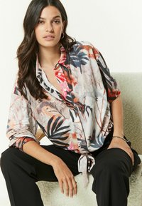 Bluse med blomstermønster i blødt materiale, der har livlige farver som pink, blå og orange, med en snørering i taljen og knaplukning foran.